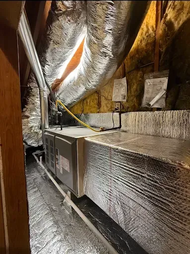 Attic HVAC unit wrapped in reflective insulation, with ducts and pipes in a narrow crawlspace