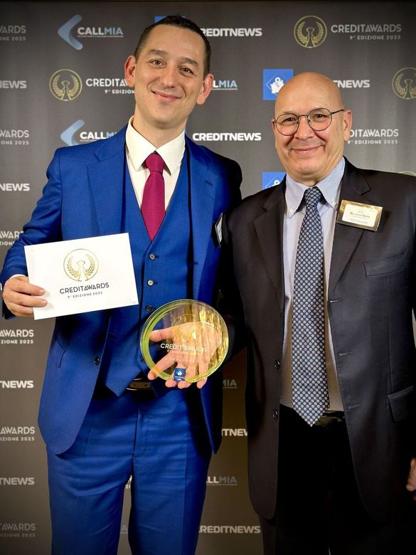 Due uomini in abito elegante posano durante una cerimonia, uno dei quali tiene in mano un premio e un certificato, sullo sfondo di un logo aziendale.