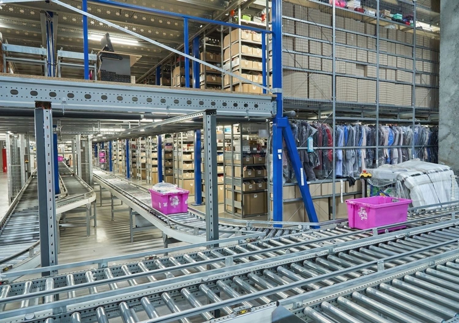 Warehouse with metal racks, conveyor rollers, and pink bins stacked with boxed inventory