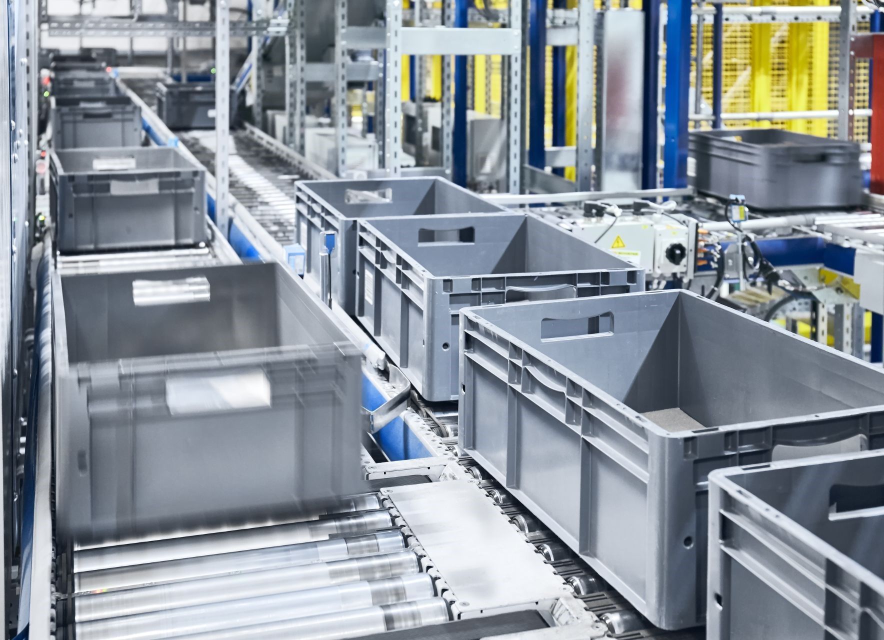 Industrial warehouse with large gray bins on a conveyor line and automated machinery in the background