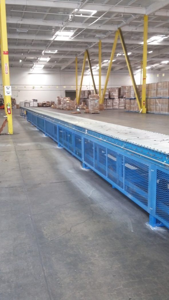 Warehouse aisle with blue safety barriers, yellow overhead supports, and stacked boxes in the background