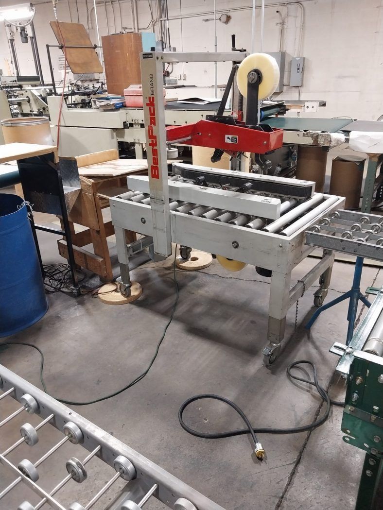Industrial conveyor and packaging machine in a workshop, with red overhead arm and metal rollers.