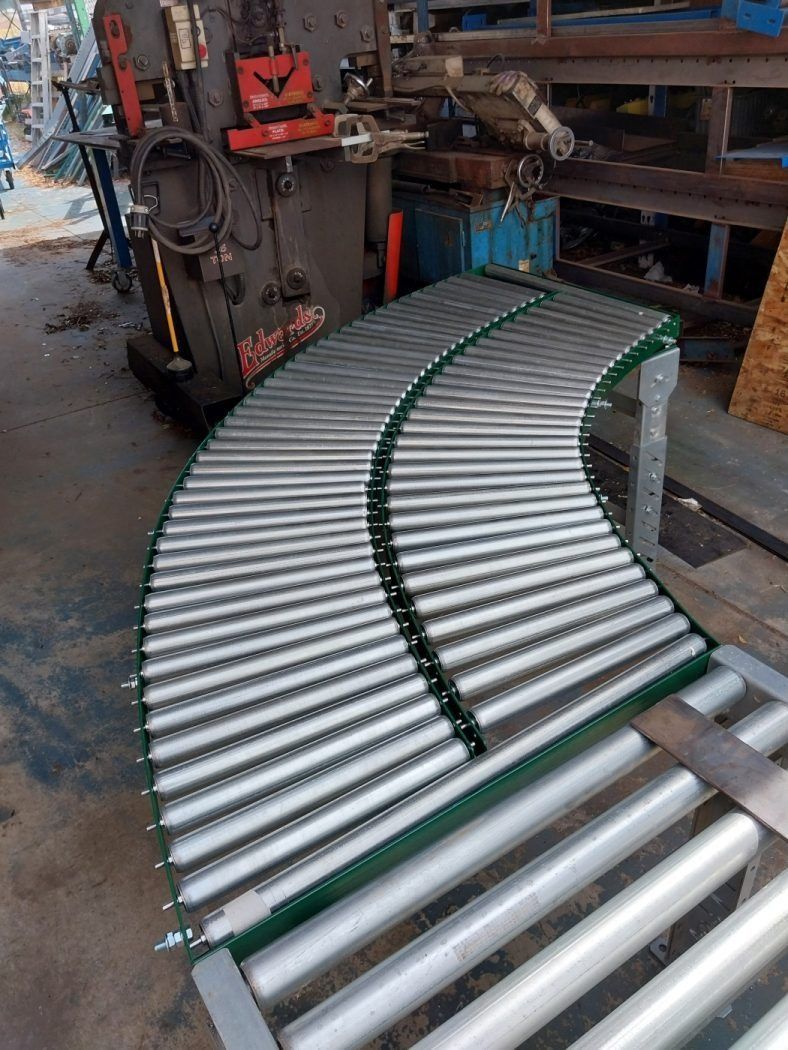 Curved metal roller conveyor in an industrial workshop, with silver rollers and green side rails