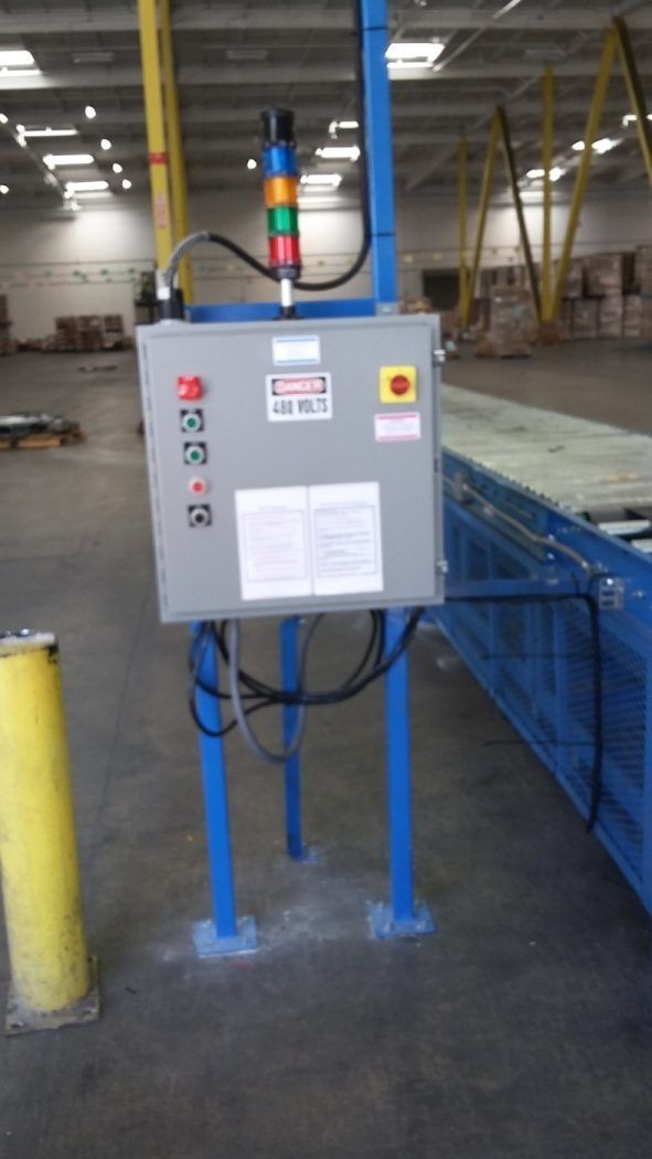 Gray industrial control box on blue legs in a factory aisle, with warning lights and cables attached
