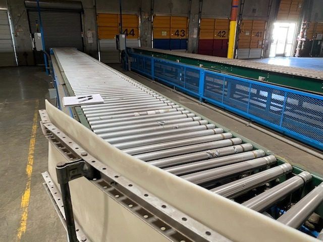 Curved conveyor rollers in an industrial warehouse with blue safety railings and stacked crates nearby
