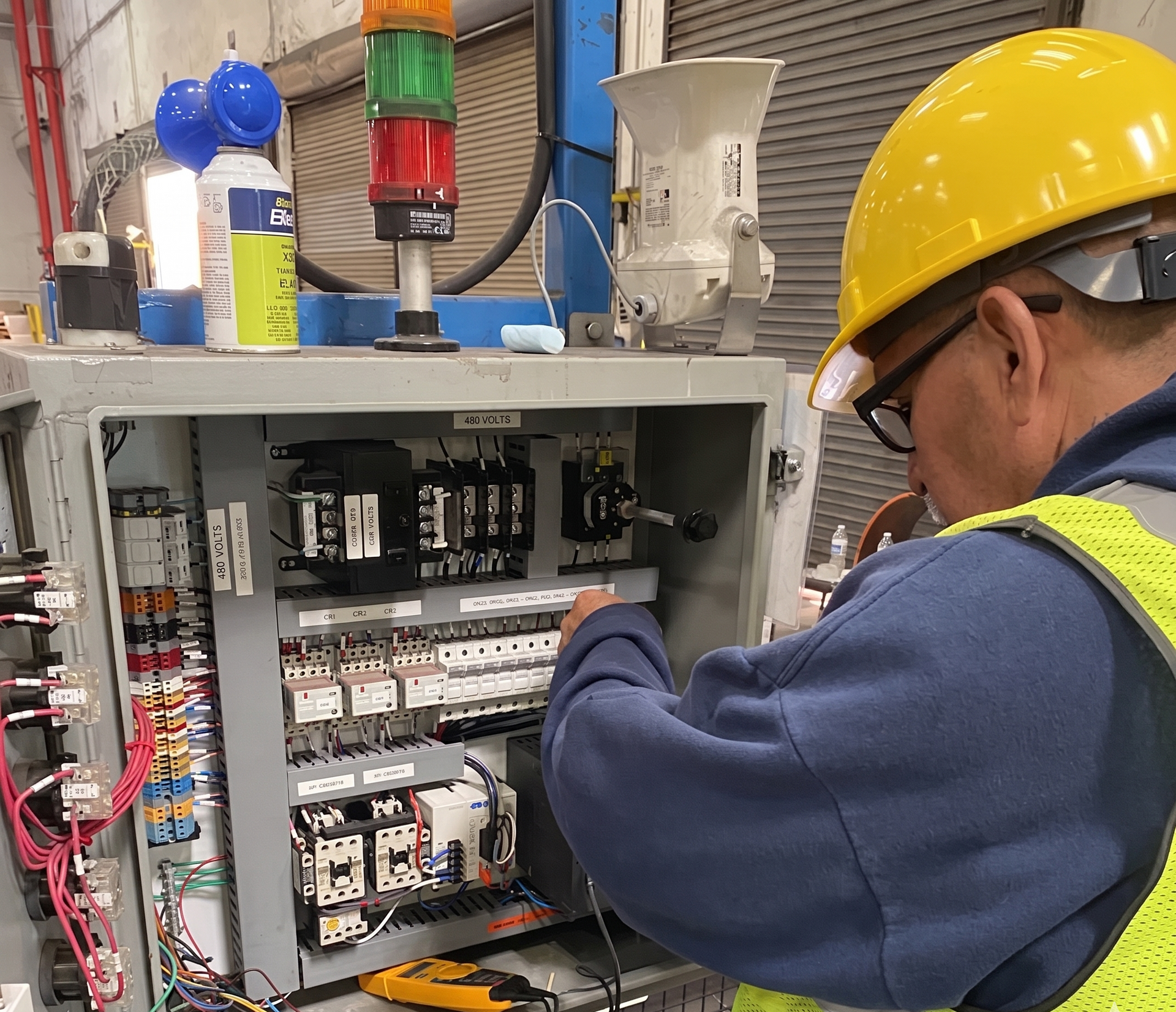 Technician in yellow hard hat inspecting an open electrical control panel with colored indicator lights.