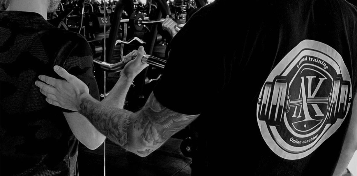 Een man helpt een andere man met gewichtheffen in een sportschool. Een van hen heeft een tatoeage op zijn arm en een logo op zijn shirt.