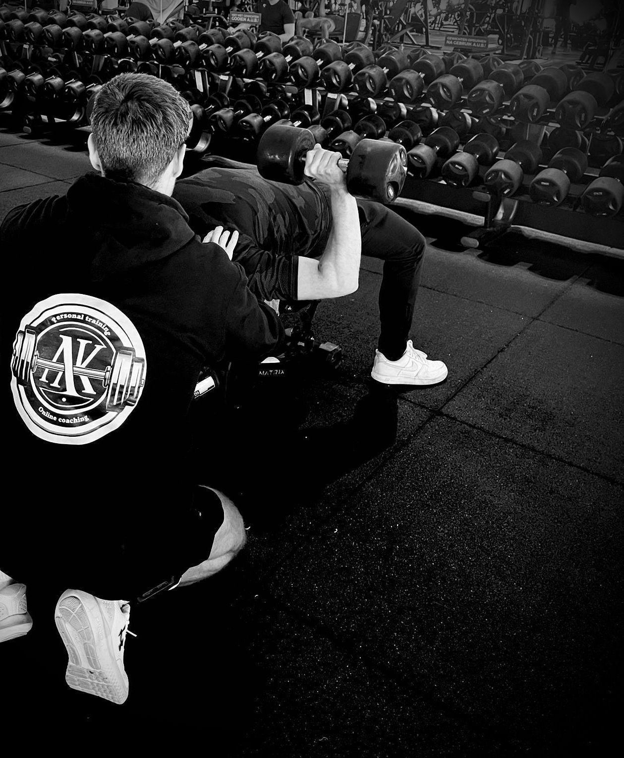 Man die halters optilt bij een bankdrukbank, met een spotter in de buurt in een sportschool. Zwart-witfoto.
