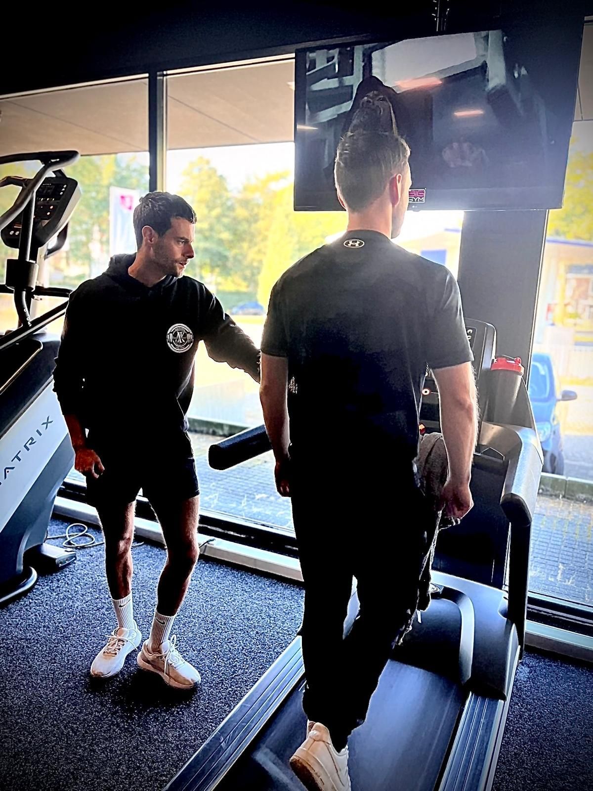 Een man helpt een andere man met gewichtheffen in een sportschool. Een van hen heeft een tatoeage op zijn arm en een logo op zijn shirt.
