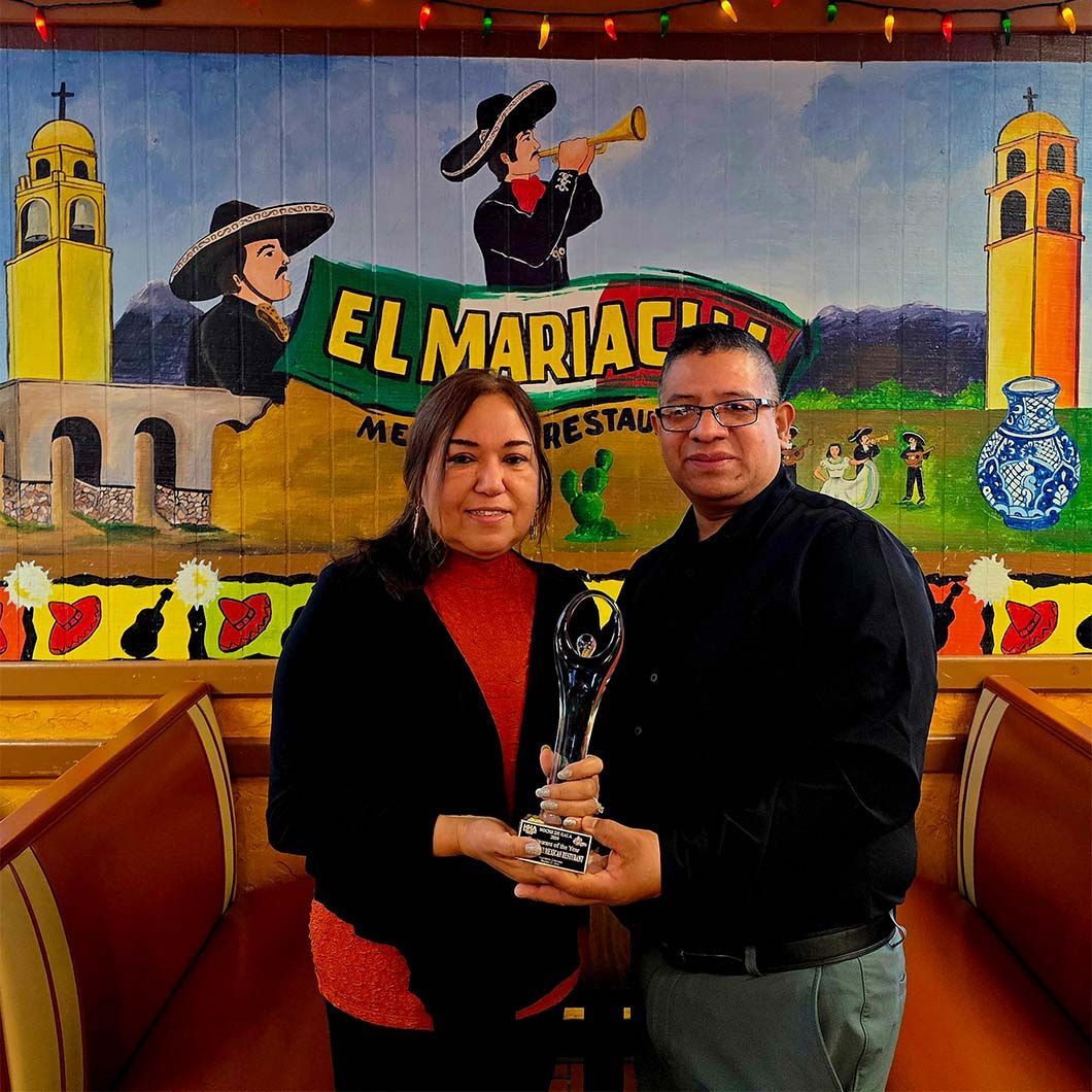Two people posing with a trophy in front of an El Mariachi restaurant mural.