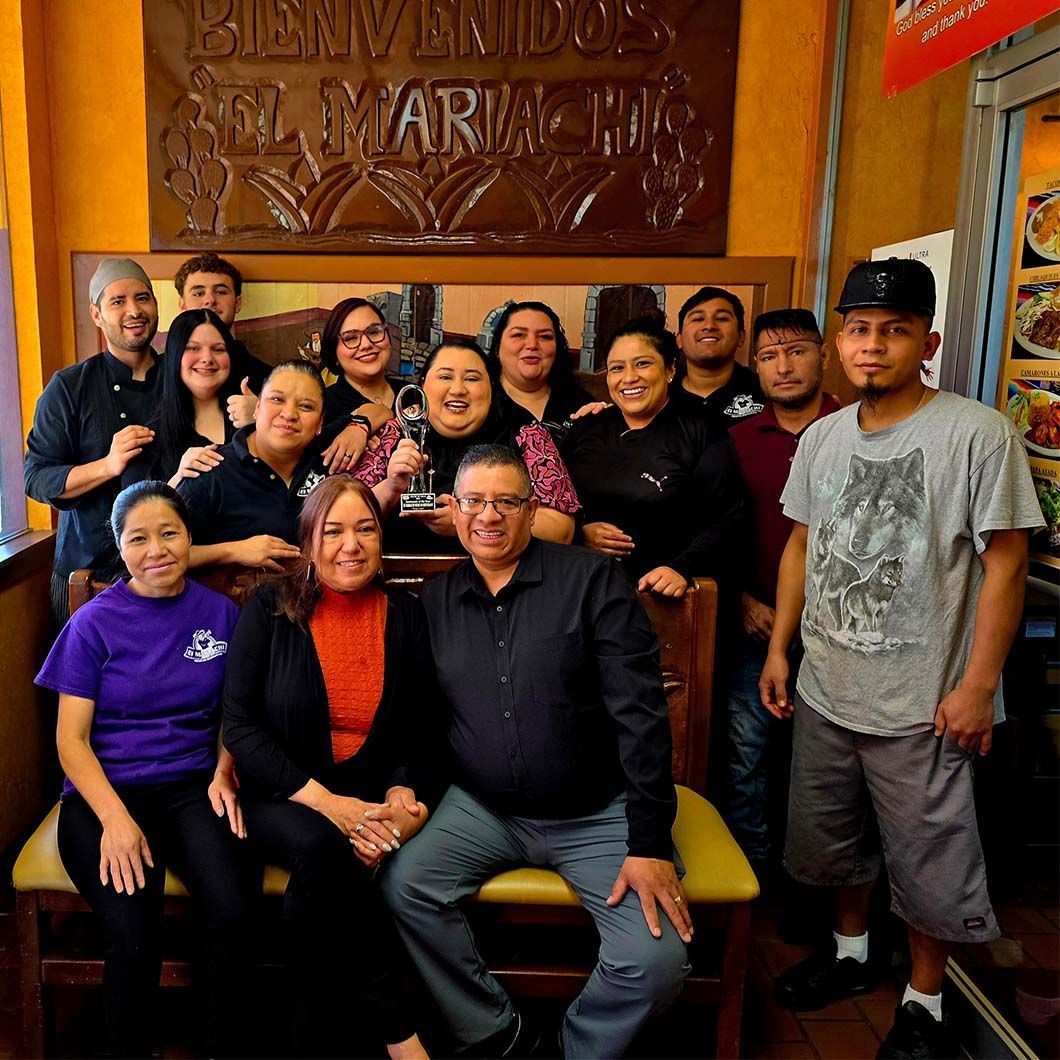 Group of smiling people posing indoors in front of a carved wooden sign.