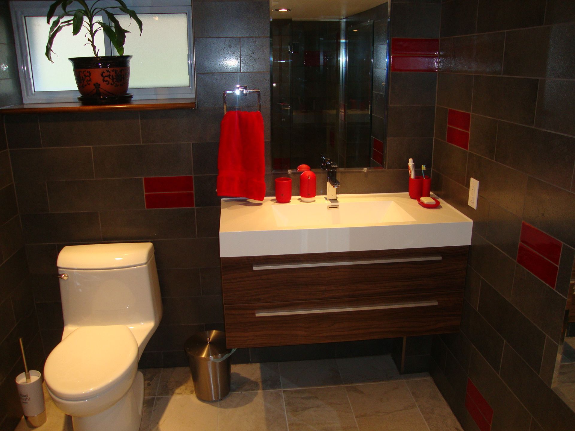 Salle de bains moderne avec murs carrelés gris foncé, meuble-lavabo en bois, lavabo blanc, touches de rouge et plante en pot.