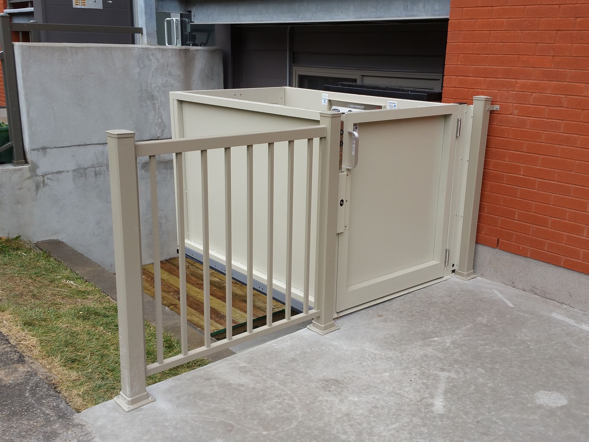 Enclos à poubelles en métal beige avec portail, à côté d'un bâtiment en briques rouges et de béton.