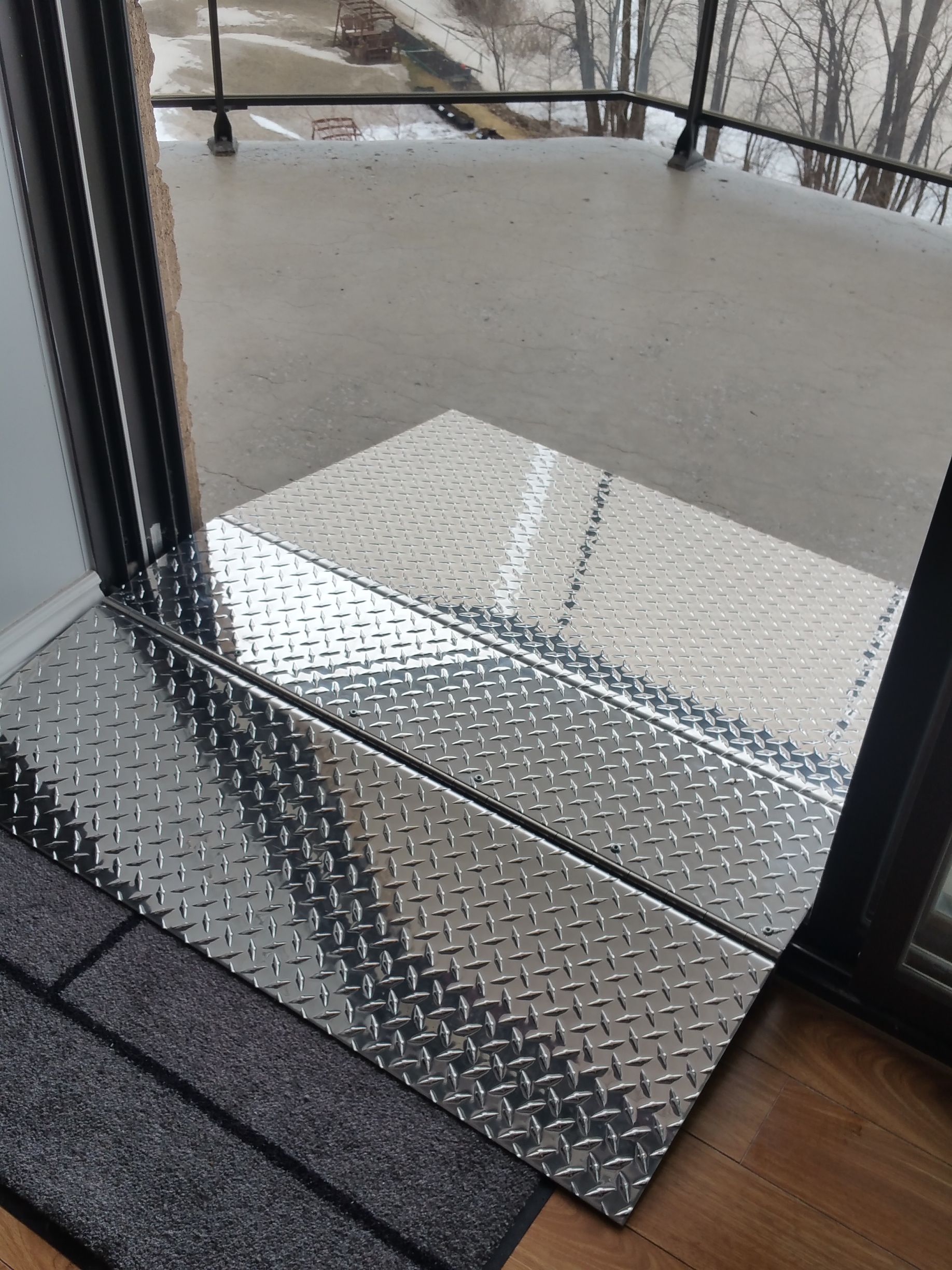 Rampe métallique à damier menant d'une porte à un balcon extérieur. Un paillasson est placé devant la porte.