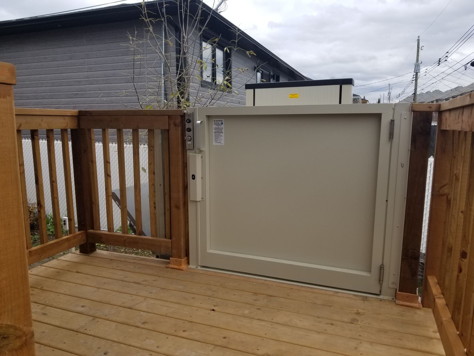 Porte d'accès pour fauteuil roulant beige sur une terrasse en bois, à côté d'une rambarde en bois, avec un bâtiment gris en arrière-plan.