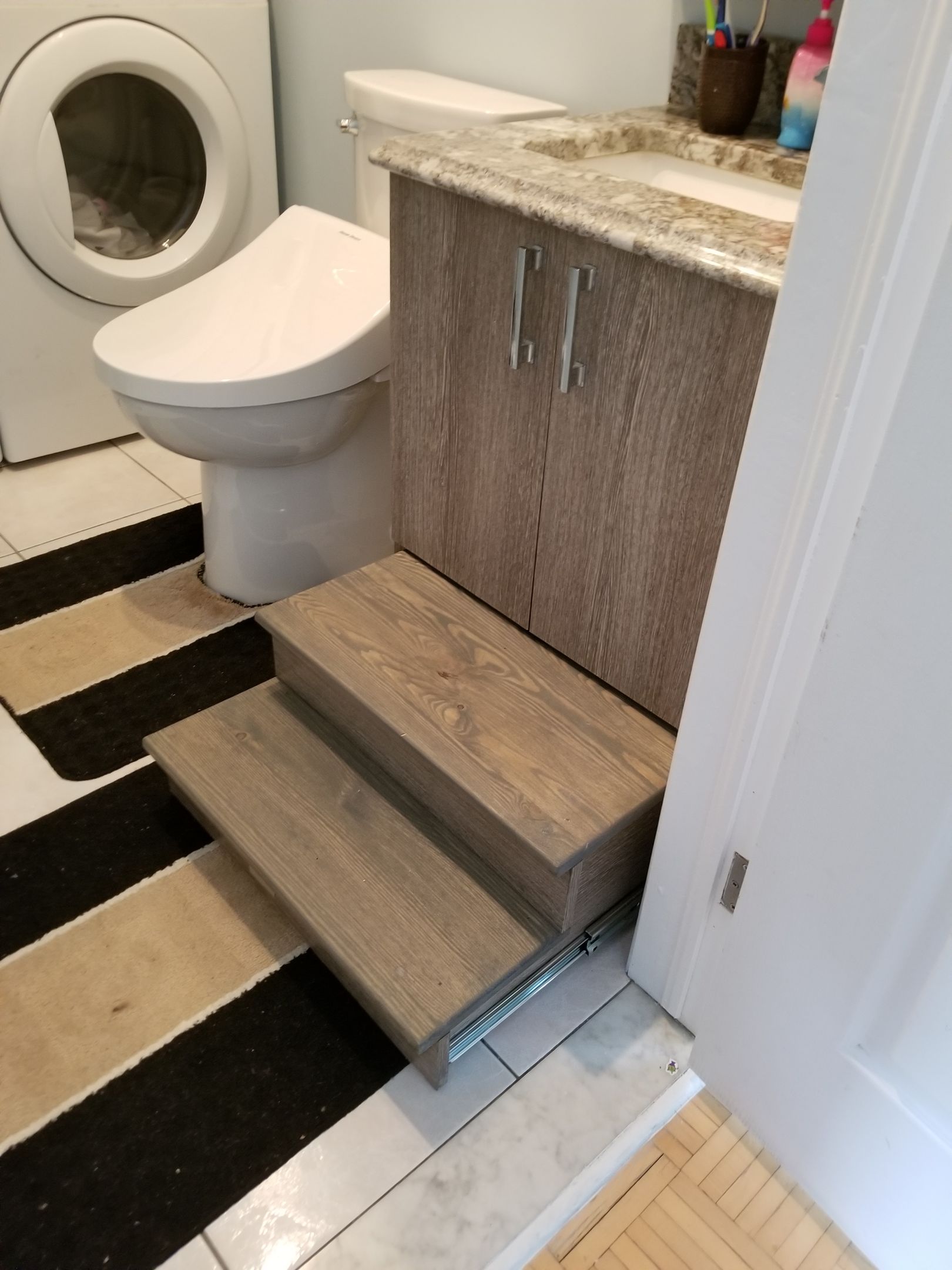 Des marches en bois dans la salle de bains mènent aux toilettes et à un lavabo ; une machine à laver est visible.