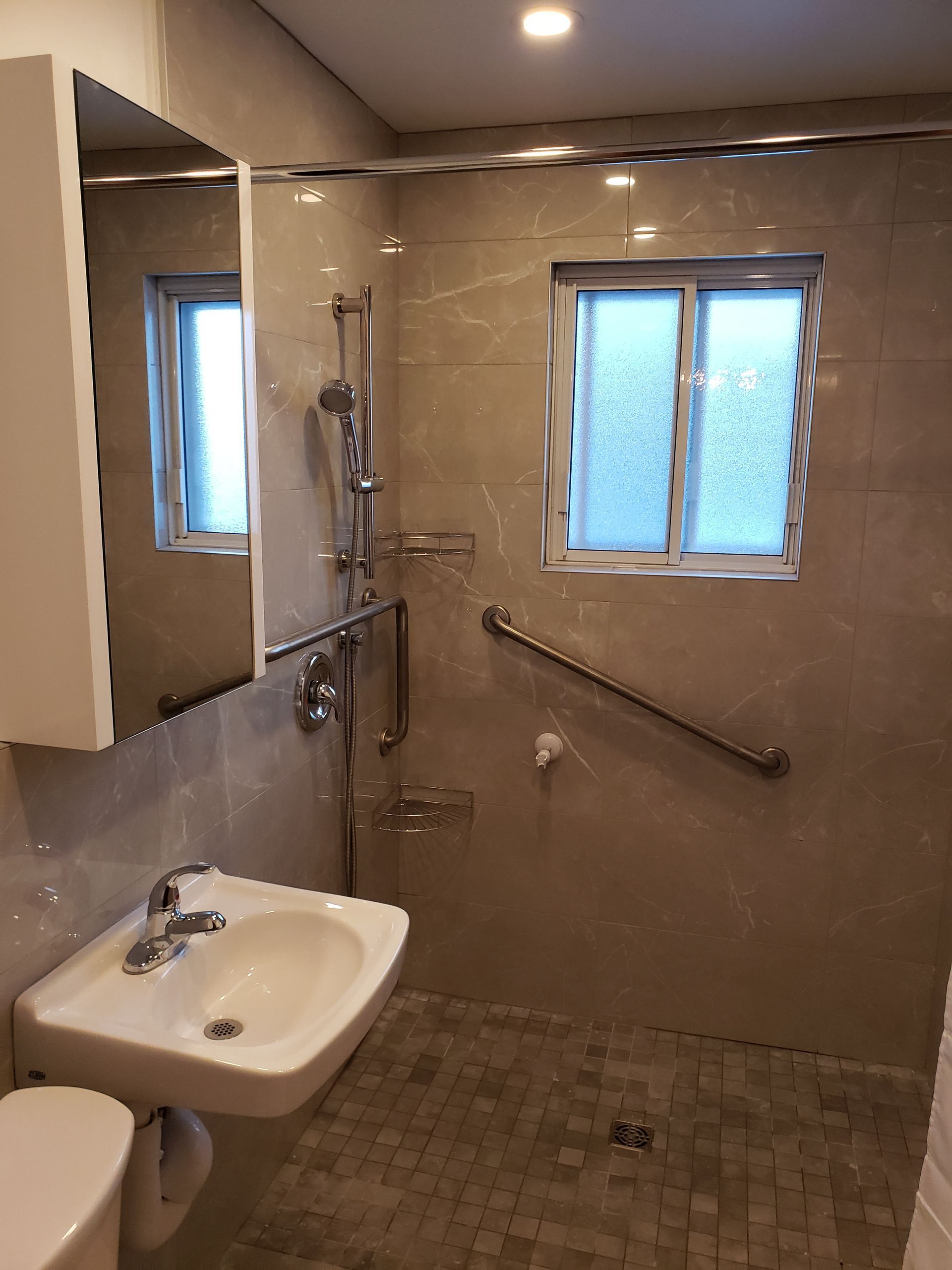 Salle de bain accessible avec douche, lavabo et barre d'appui. Carrelage beige, petite fenêtre et accessoires muraux.