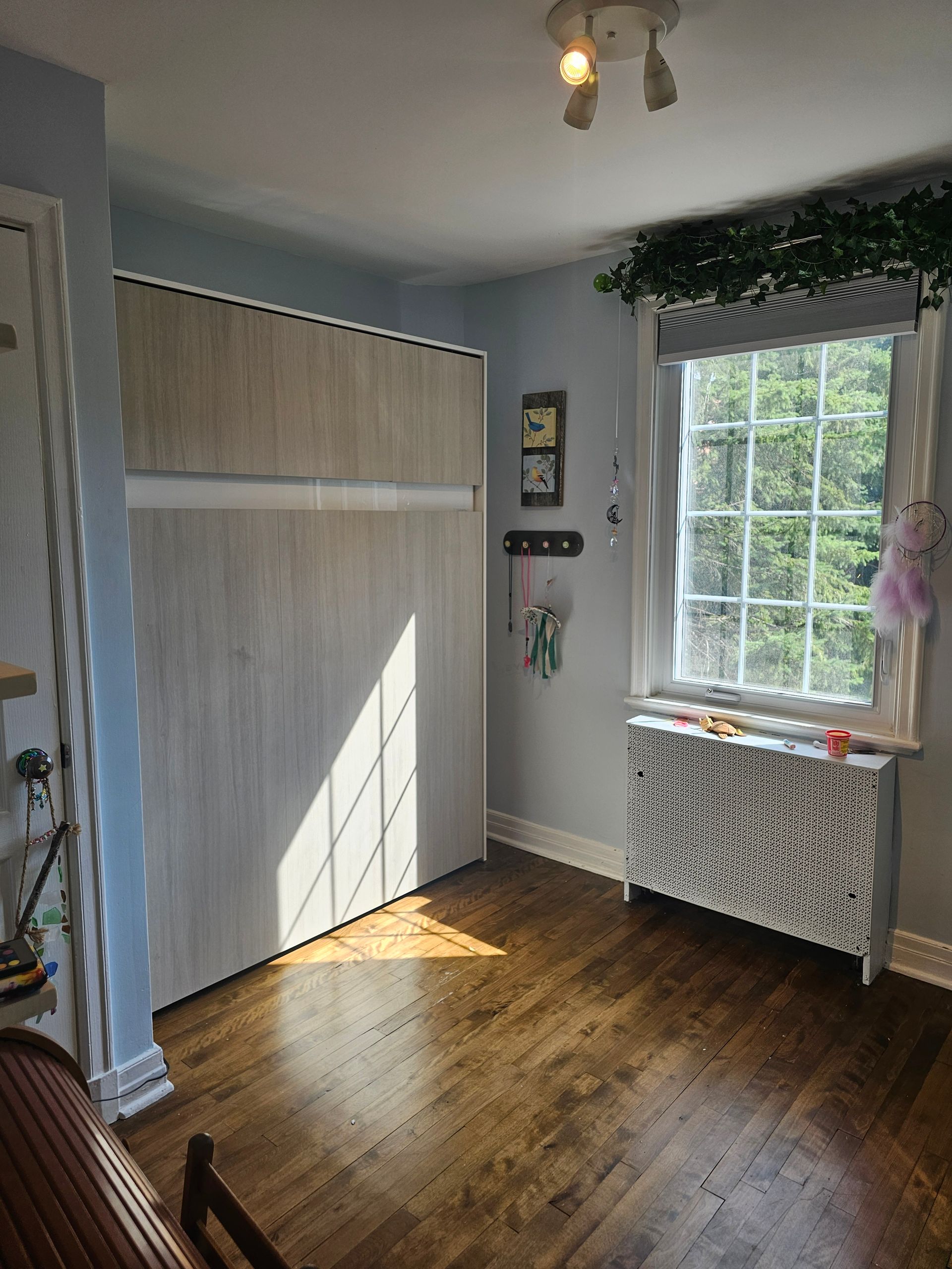 Chambre avec parquet, murs bleu clair, une armoire fermée, une fenêtre avec de la verdure et un radiateur.