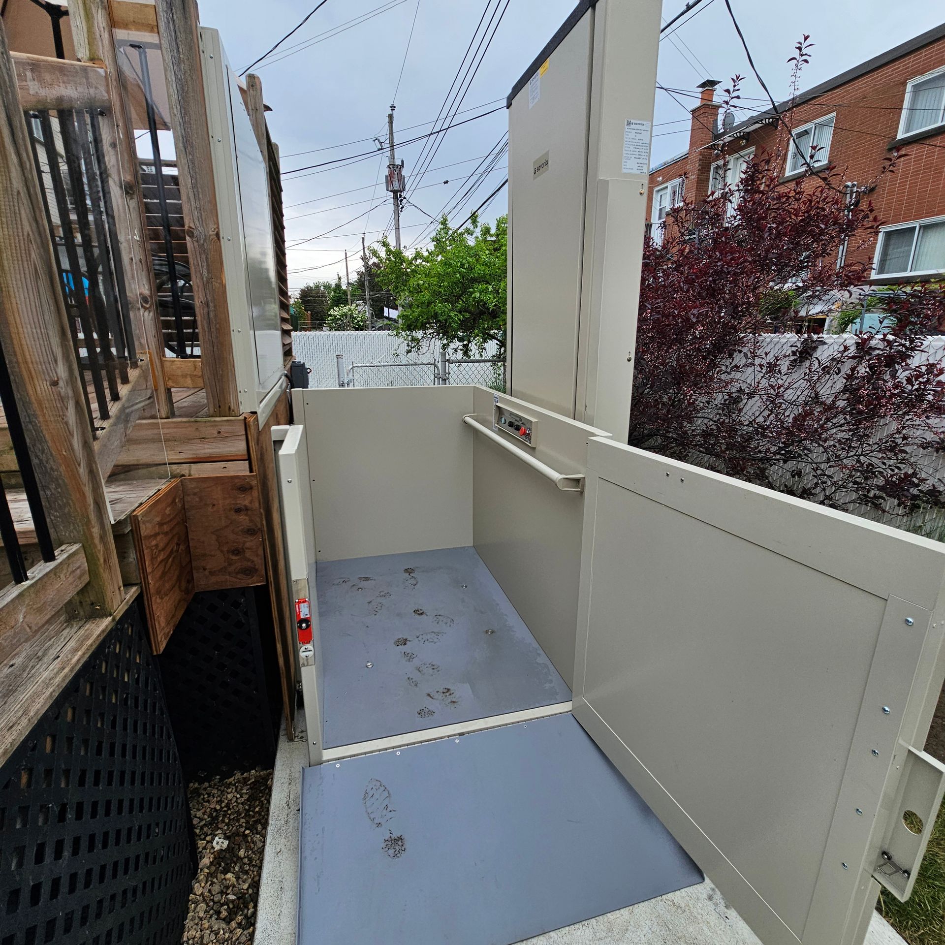 Plateforme élévatrice extérieure beige pour fauteuils roulants, à côté d'escaliers en bois et d'un bâtiment en briques.