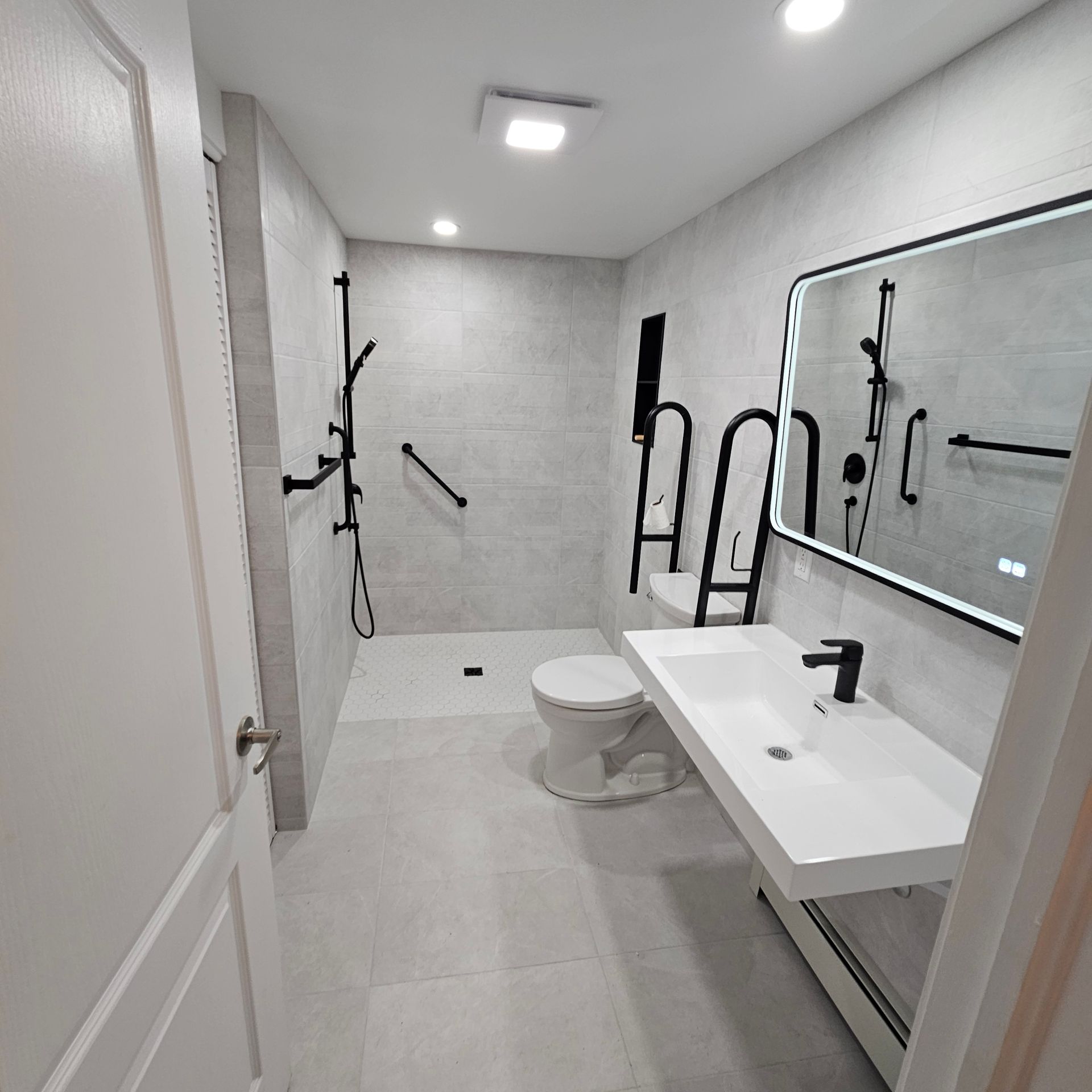 Salle de bains moderne et accessible avec robinetterie noire, douche, toilettes et meuble-lavabo. Carrelage gris partout.