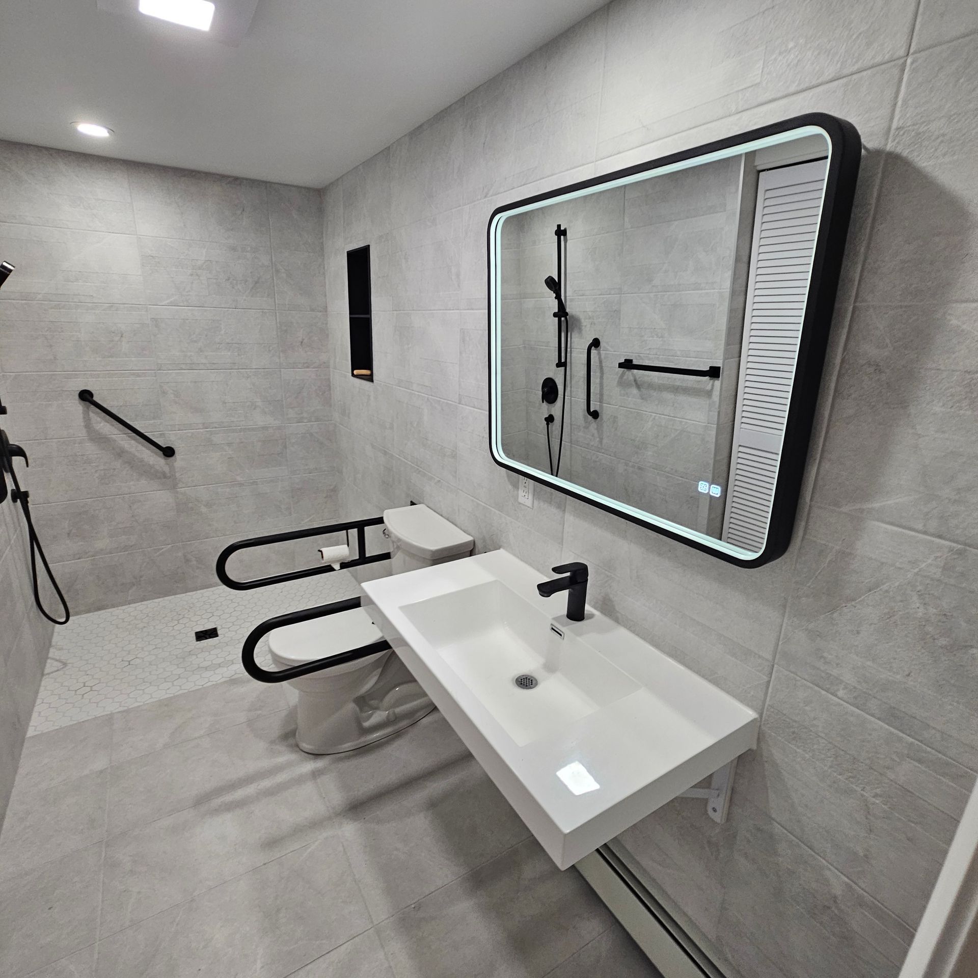 Salle de bain accessible : carrelage gris, lavabo blanc, robinetterie noire, barres d’appui, toilettes avec barres de soutien et grand miroir.