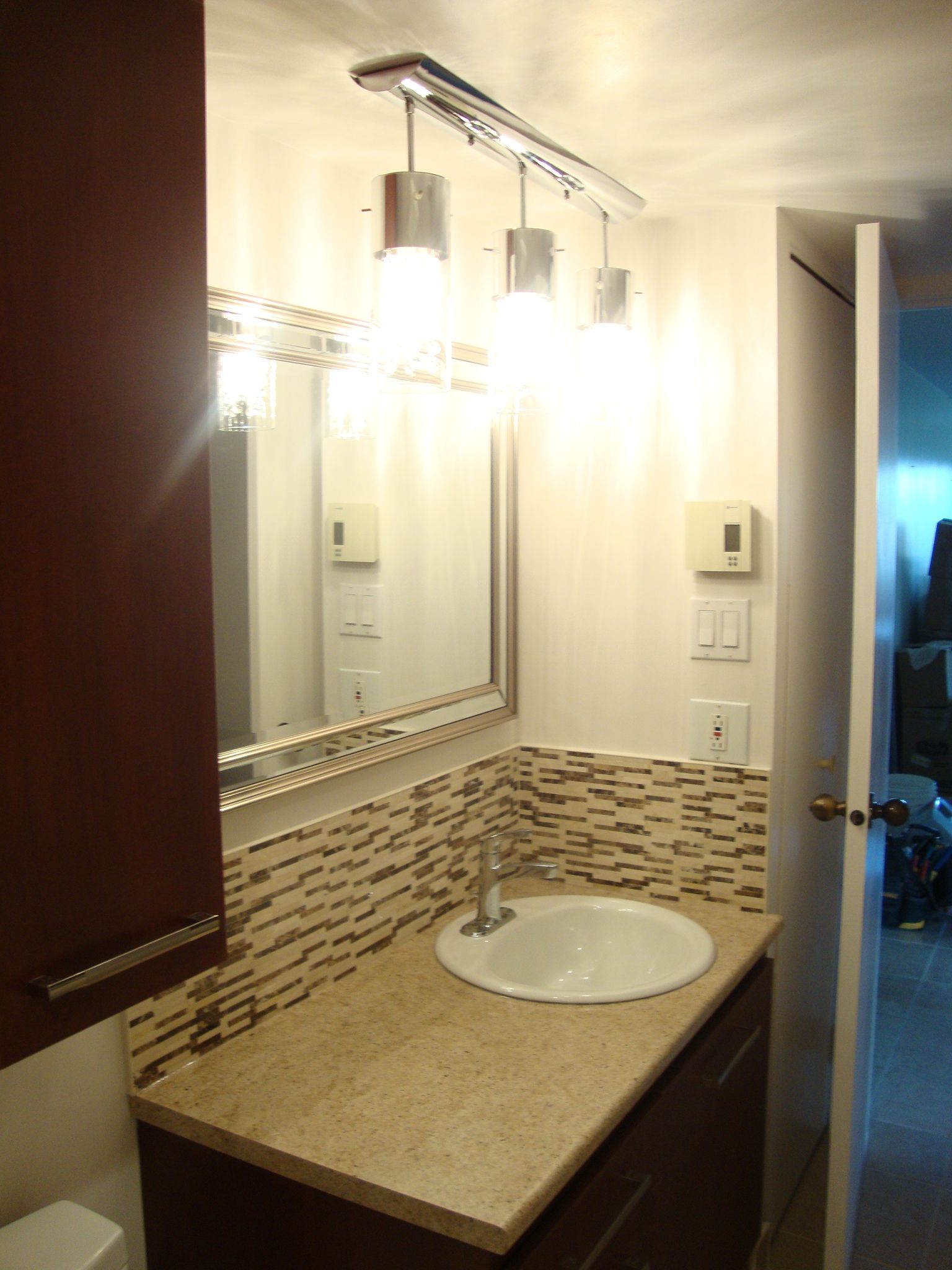 Meuble-lavabo avec lavabo, miroir et luminaire. Armoires brun foncé, comptoir beige et crédence en mosaïque.