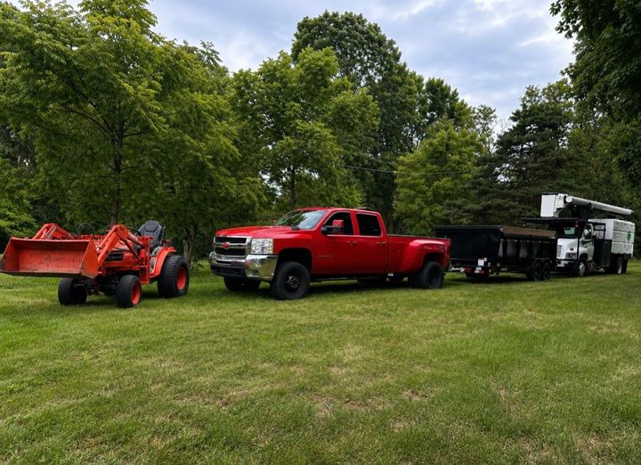 Orange tractor, red pickup truck, and black trailer parked on a grassy field with trees in the background