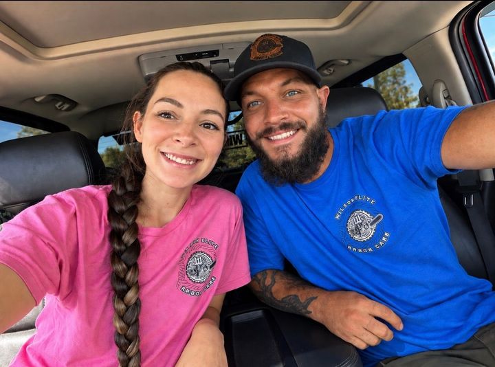 Two smiling people seated in a car, wearing pink and blue shirts, taking a selfie