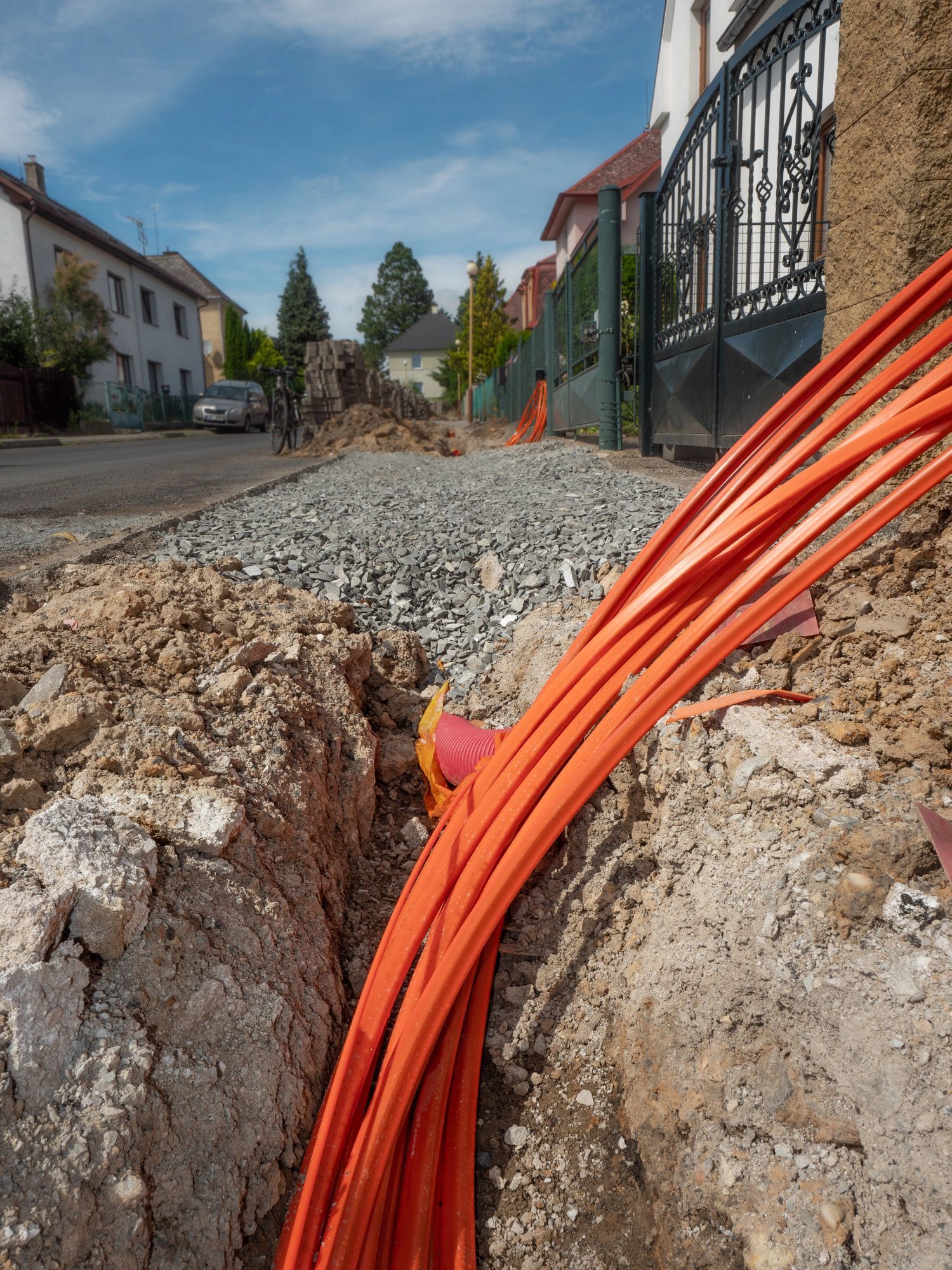 Trincea con cavi in fibra ottica arancioni. Strada in costruzione; case e cancello sullo sfondo.