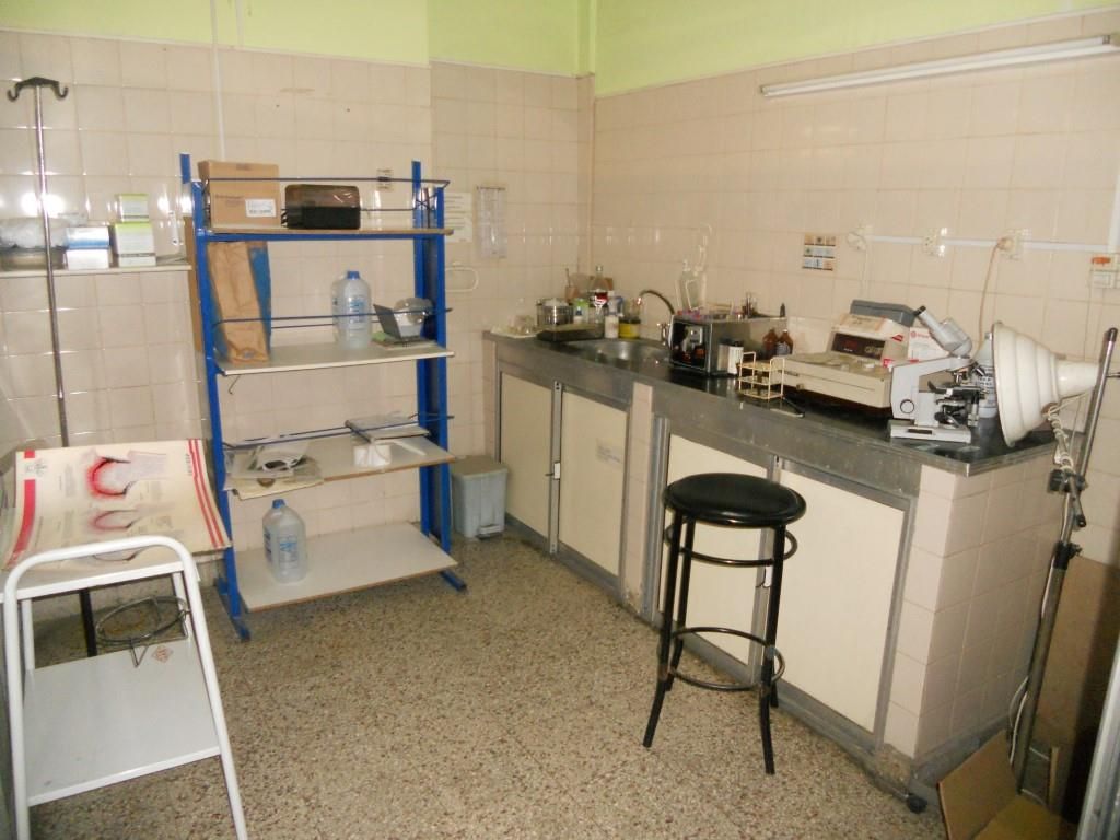 Pequeño cuarto de servicio con estantes, armarios, fregadero, taburetes y artículos de limpieza sobre suelo de baldosas.