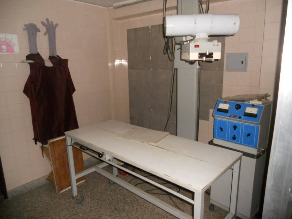 Sala de exploración con camilla, dispositivo médico montado en la pared y armario azul para equipos.