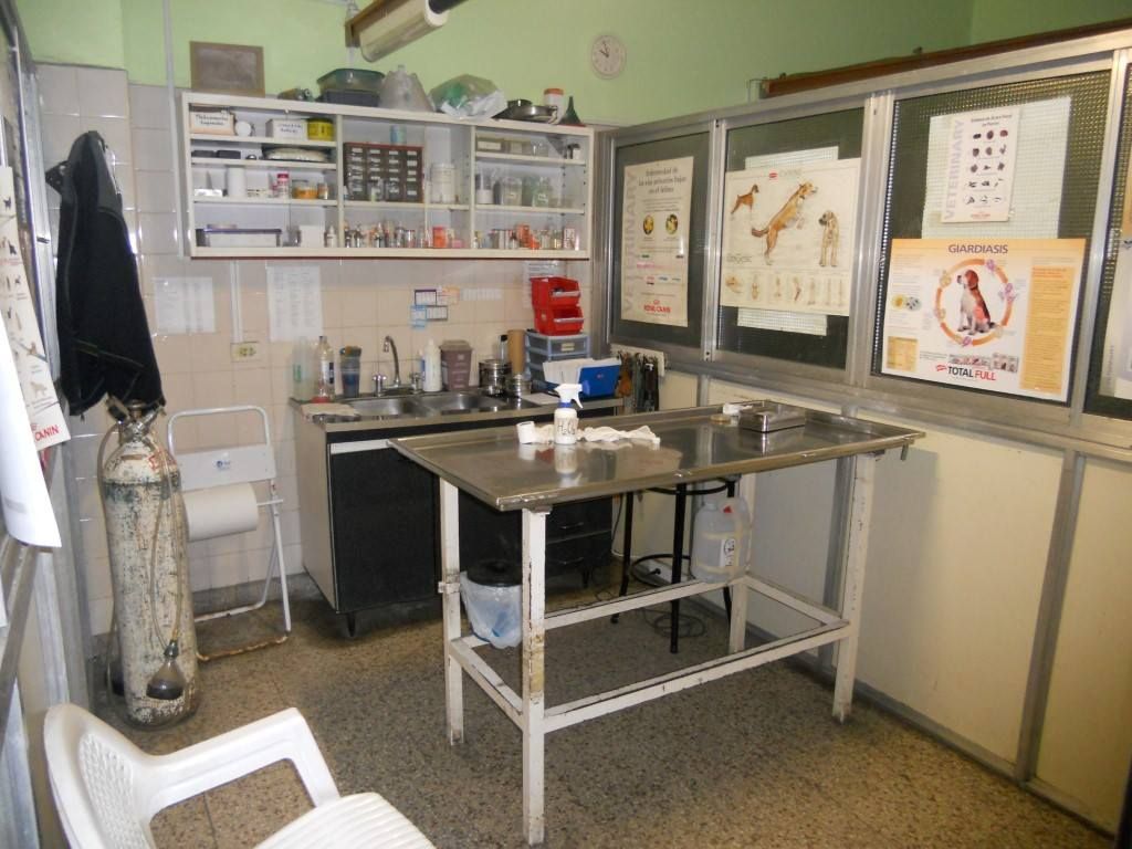 Pequeña sala de exploración clínica con camilla metálica, armarios, lavabo y láminas explicativas en la pared.