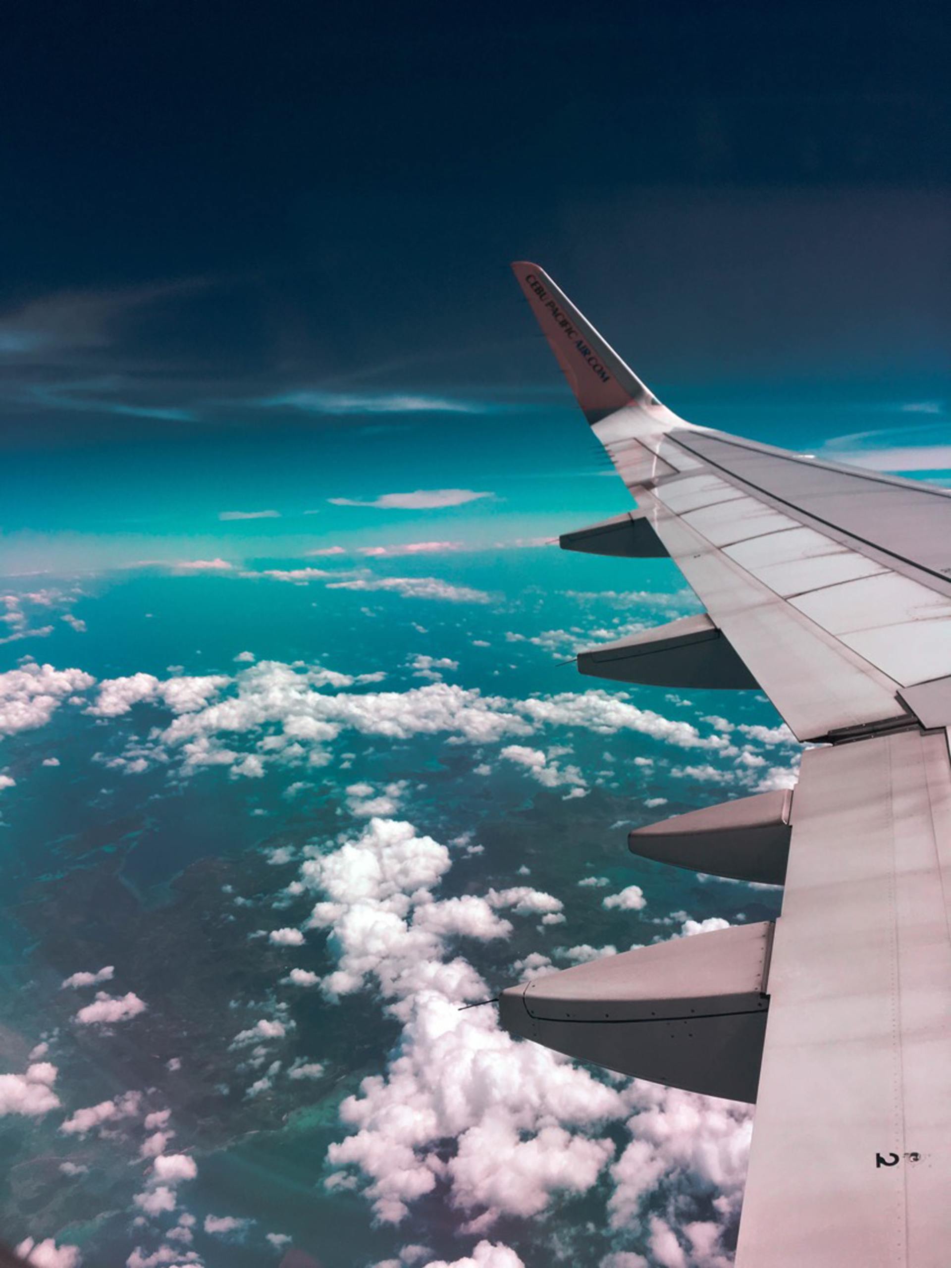 Airplane wing soaring above fluffy white clouds under a turquoise and dark blue sky.