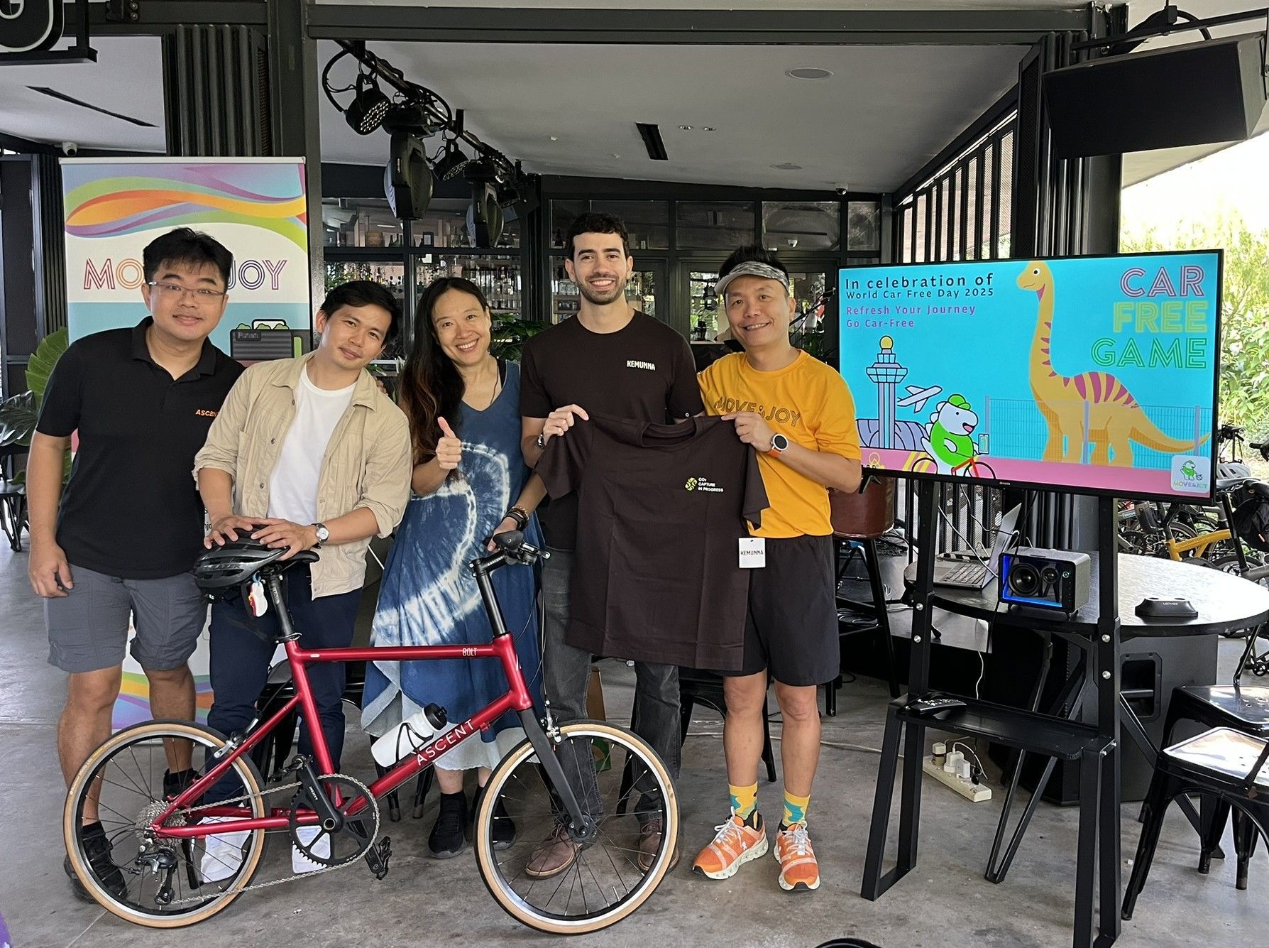 Five people posing with a red bicycle beside a colorful display screen indoors