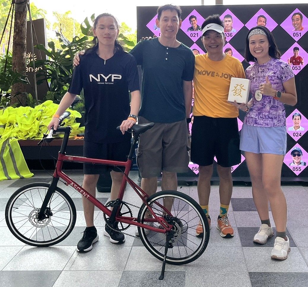 Four people pose outdoors; one holds a red bicycle in front of a decorative event backdrop.
