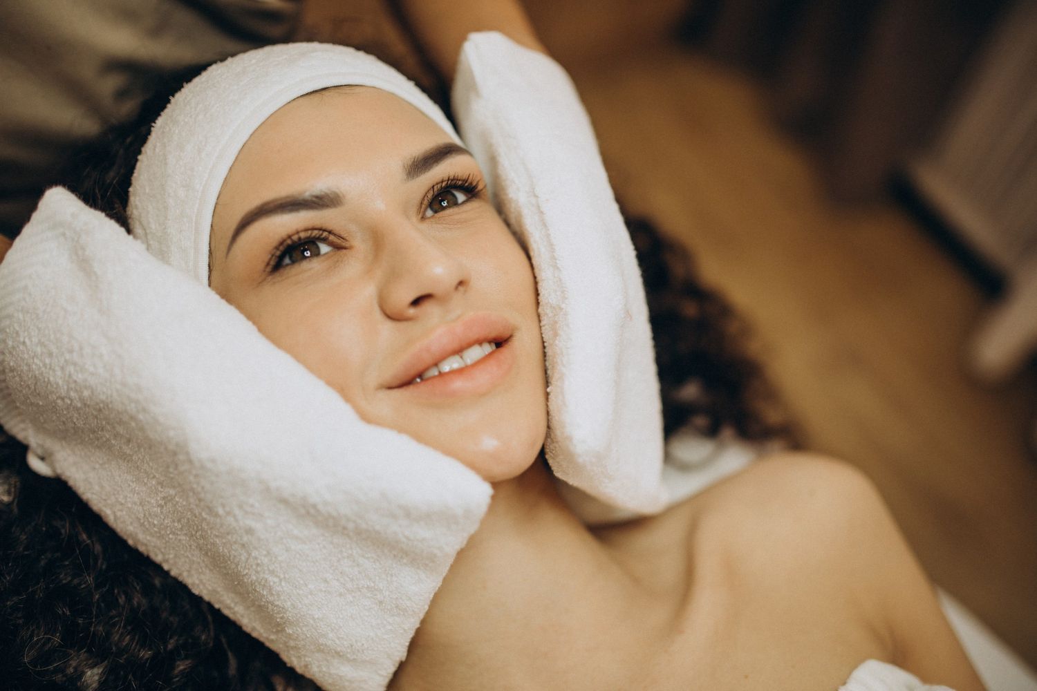 A person with a spa headband lies back with white towels placed against each side of their face.