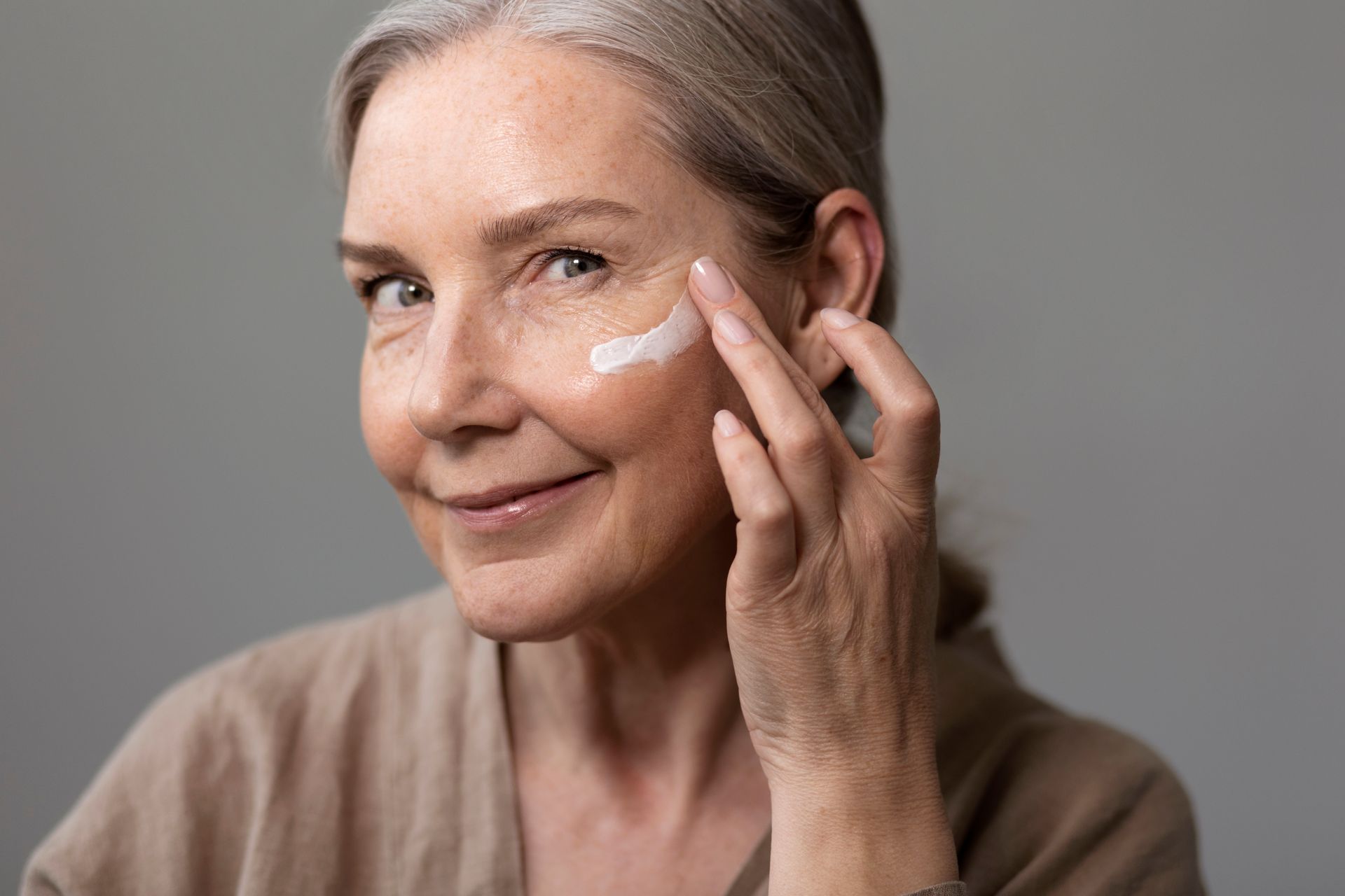 Person applying cream to cheek in a close-up portrait, wearing a beige top against a gray background