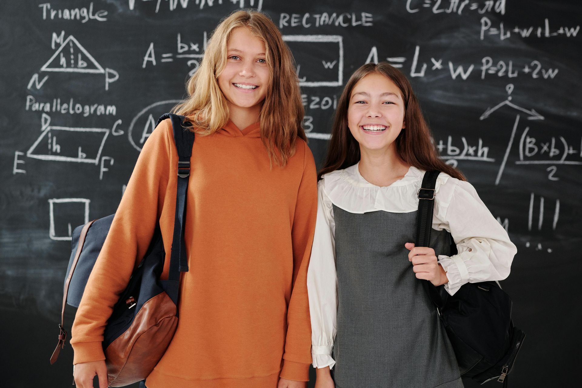 Two smiling girls with backpacks in front of a chalkboard covered in math formulas.