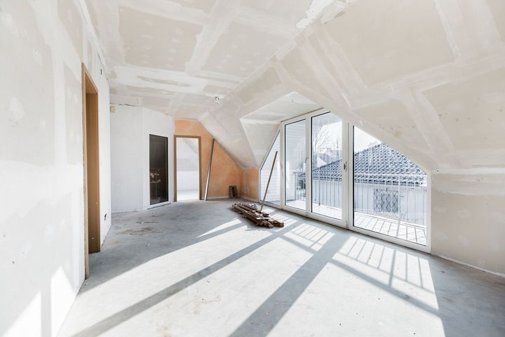 Bright unfinished attic room with white drywall, large windows, and sunlight casting shadows on the floor.