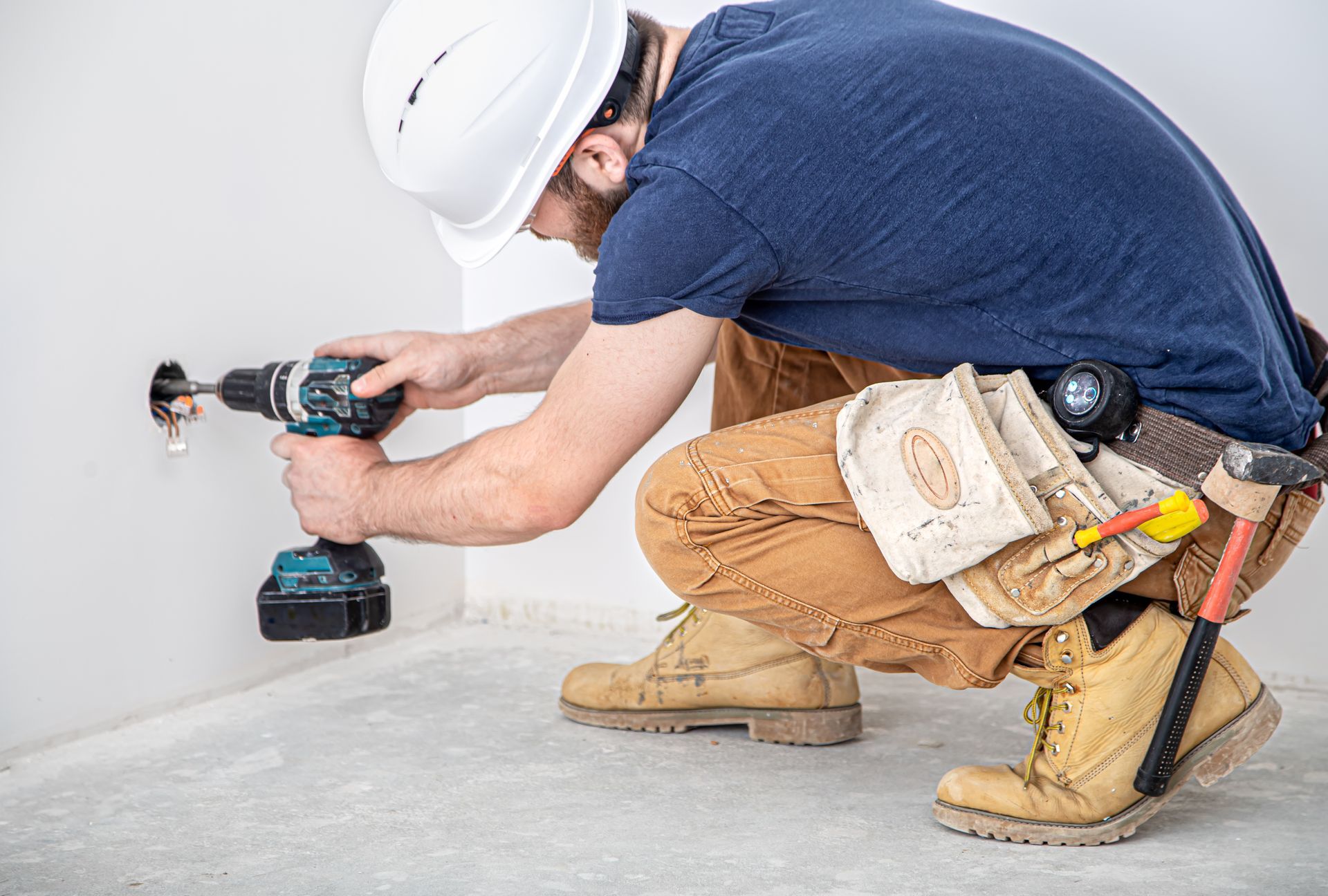 Worker in white hard hat drilling into a wall while kneeling on a floor with tools and a tool belt nearby