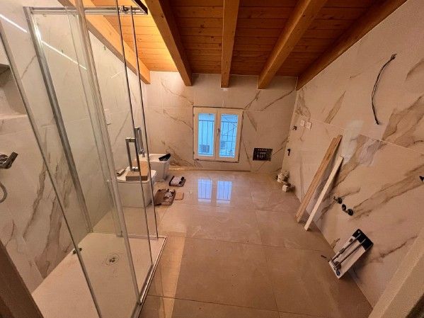 Bagno con pareti in marmo beige, doccia, travi in legno e finestra.