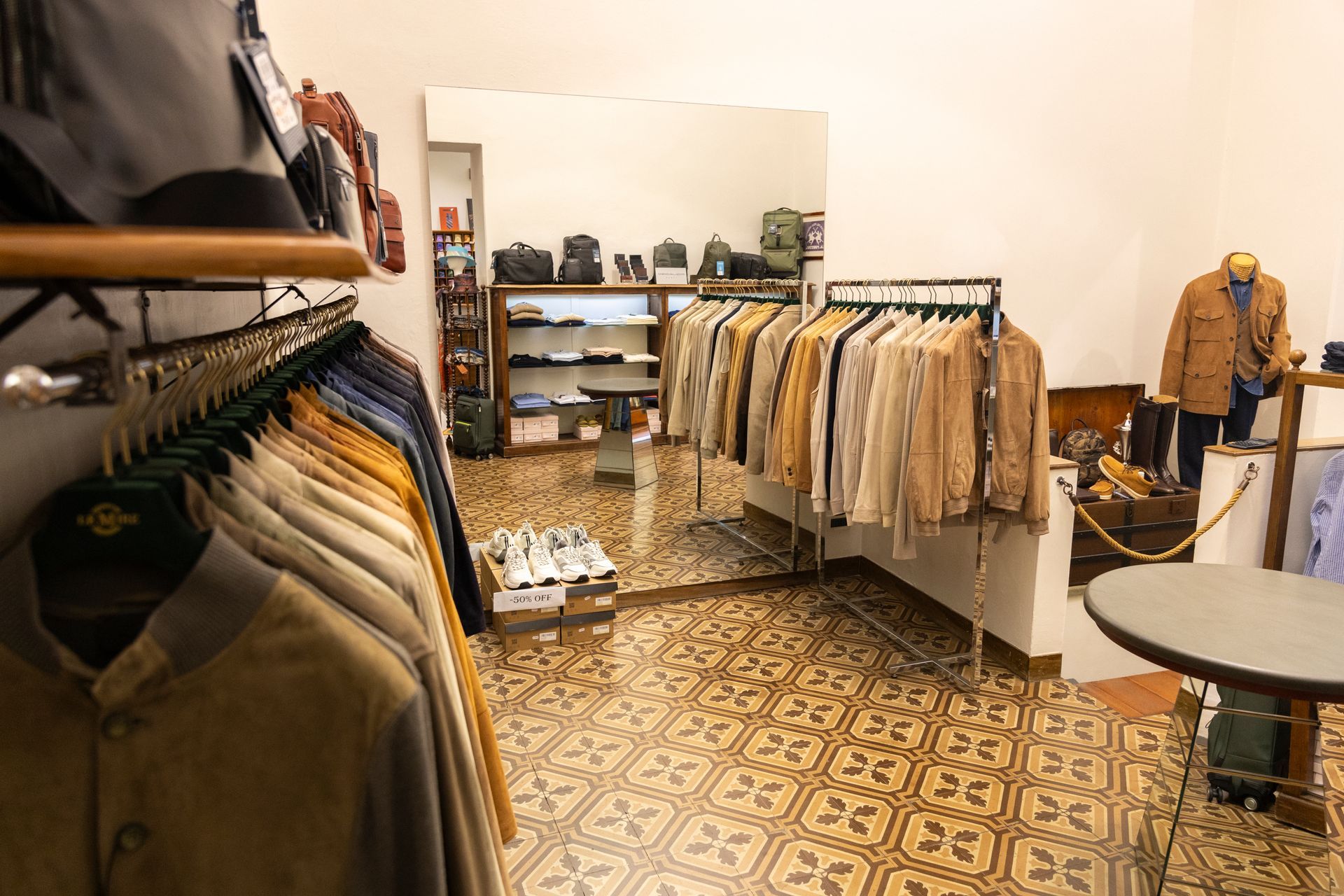 Boutique di abbigliamento con appendiabiti per giacche, specchi, scaffali e pavimento piastrellato con motivi decorativi.