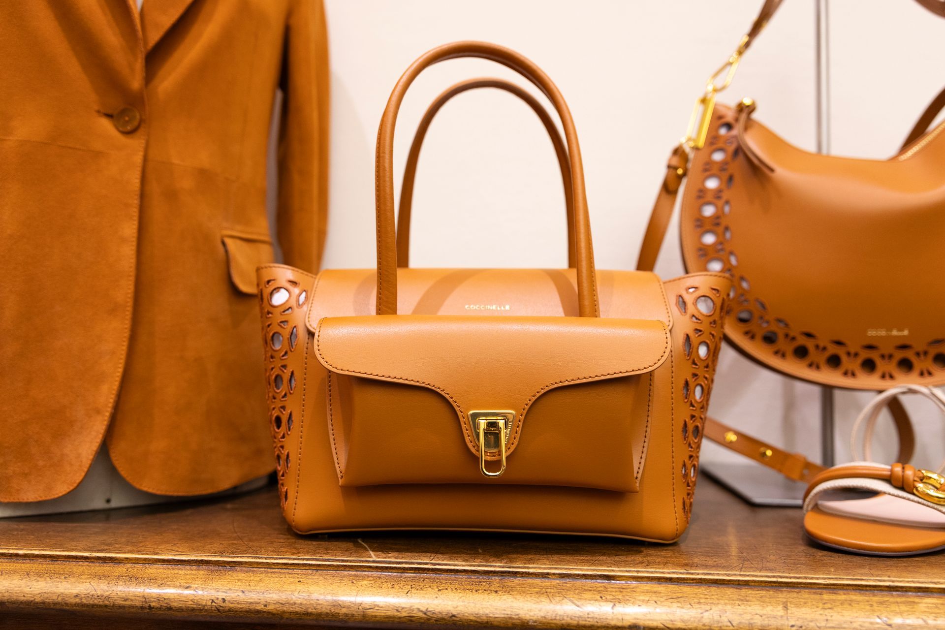 Borsa a mano in pelle color cuoio e portafoglio abbinato esposti su uno scaffale insieme a una giacca e scarpe.