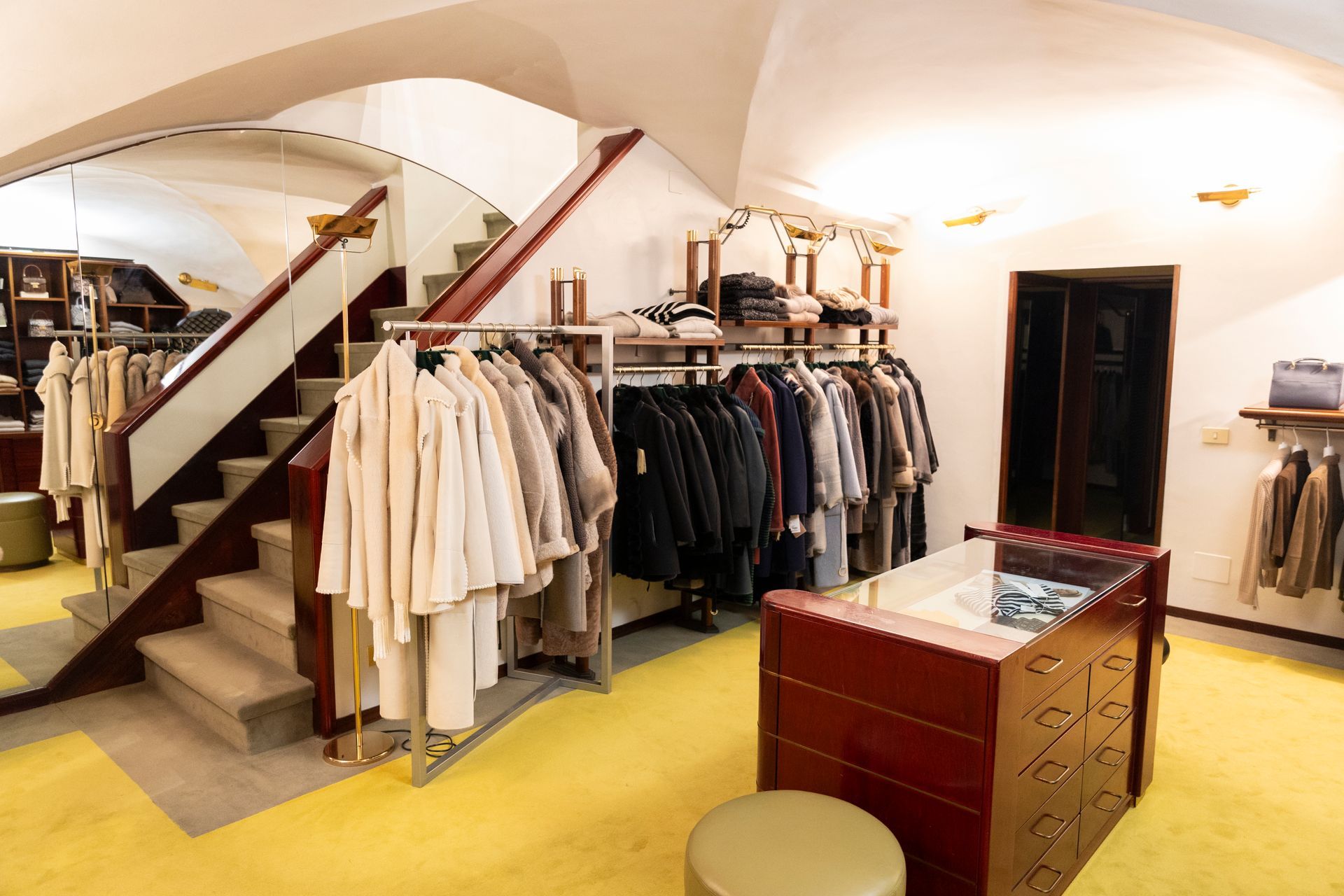 Interno di una boutique di abbigliamento con appendiabiti, scala e bancone espositivo in legno.