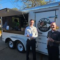 Two men stand beside a branded food truck in a sunny parking lot.