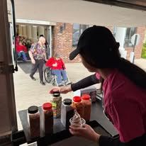 Person in a black cap serving condiments from a table to people entering a building