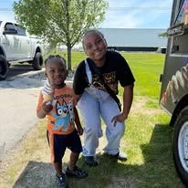 Two children crouch smiling on a grassy driveway beside parked vehicles.