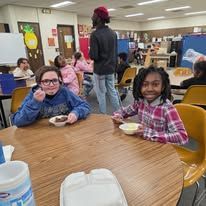 Children seated at a classroom table eating snacks, with a teacher nearby in a colorful room