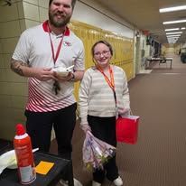 Two people standing in a school hallway, smiling, with a table and red bottle in the foreground
