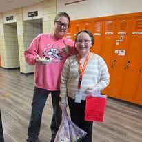 Two people smiling in a school hallway with orange lockers, one holding a red bag and the other a gift bag